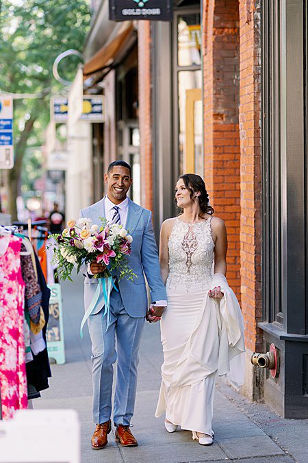 groom holding a bouquet walking with a bride down the sidewalk
