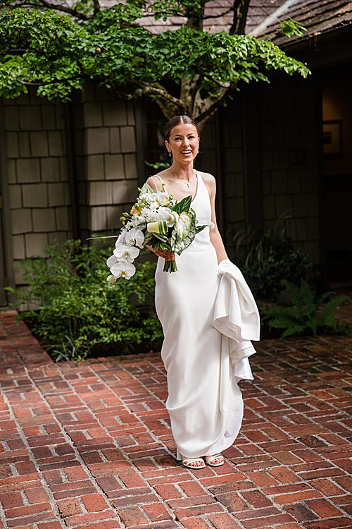 a bride stands on a brick walkway holding the bottom of her dress in one hand and her bouquet in the other hand