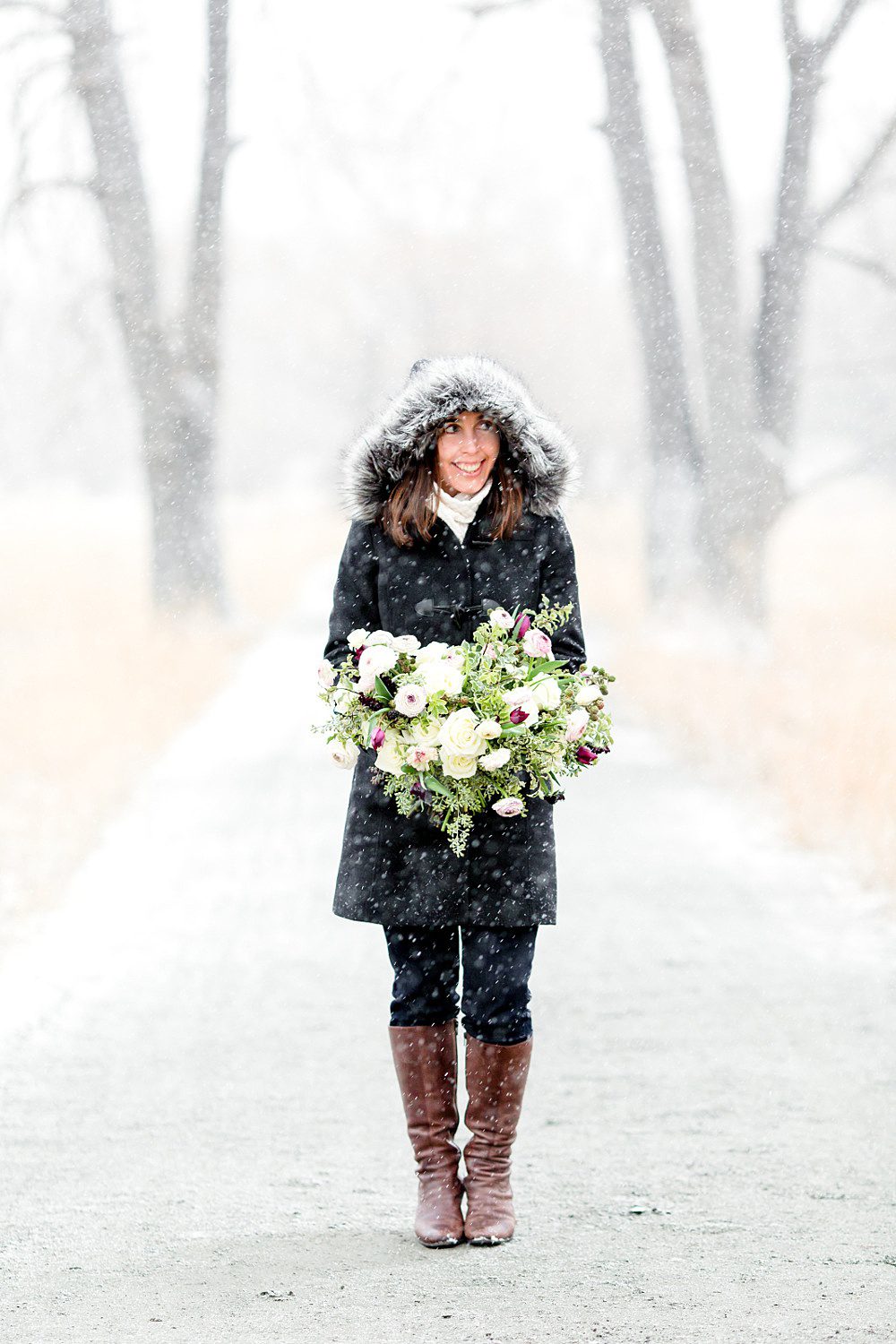 a woman wearing a coat with a furry hood holds a flower bouquet in the snow