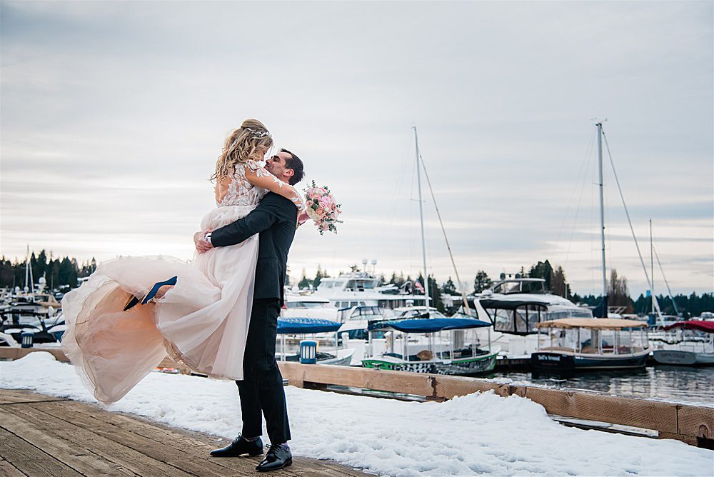 a bride and groom embrace by a frozen lake with sailboats