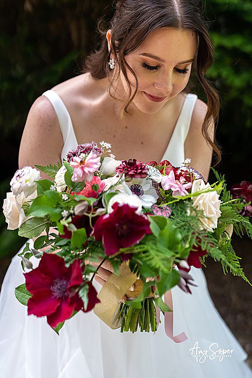 a bride leaning over and holding a bouquet with purple ranunculus and red clematis